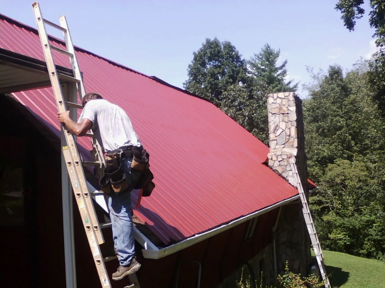 Roofer on a ladder beside a red metal roof and stone chimney — Soffit & Fascia Repair by Roofing Brothers in Scipio